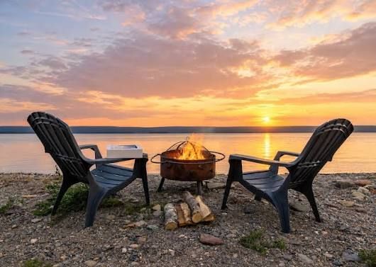 Two chairs around a fire pit at sunset on a rocky beach, with calm water and pink clouds.