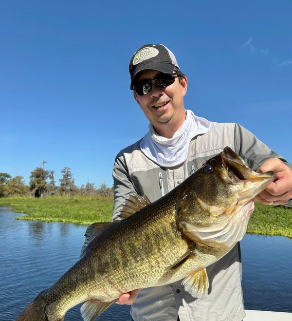 Man holding a large largemouth bass on a sunny lake, wearing a cap and sunglasses.