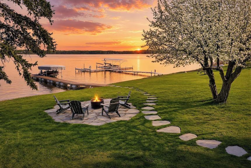Lakeside fire pit at sunset with chairs, stepping stones, docked boats, and blooming trees