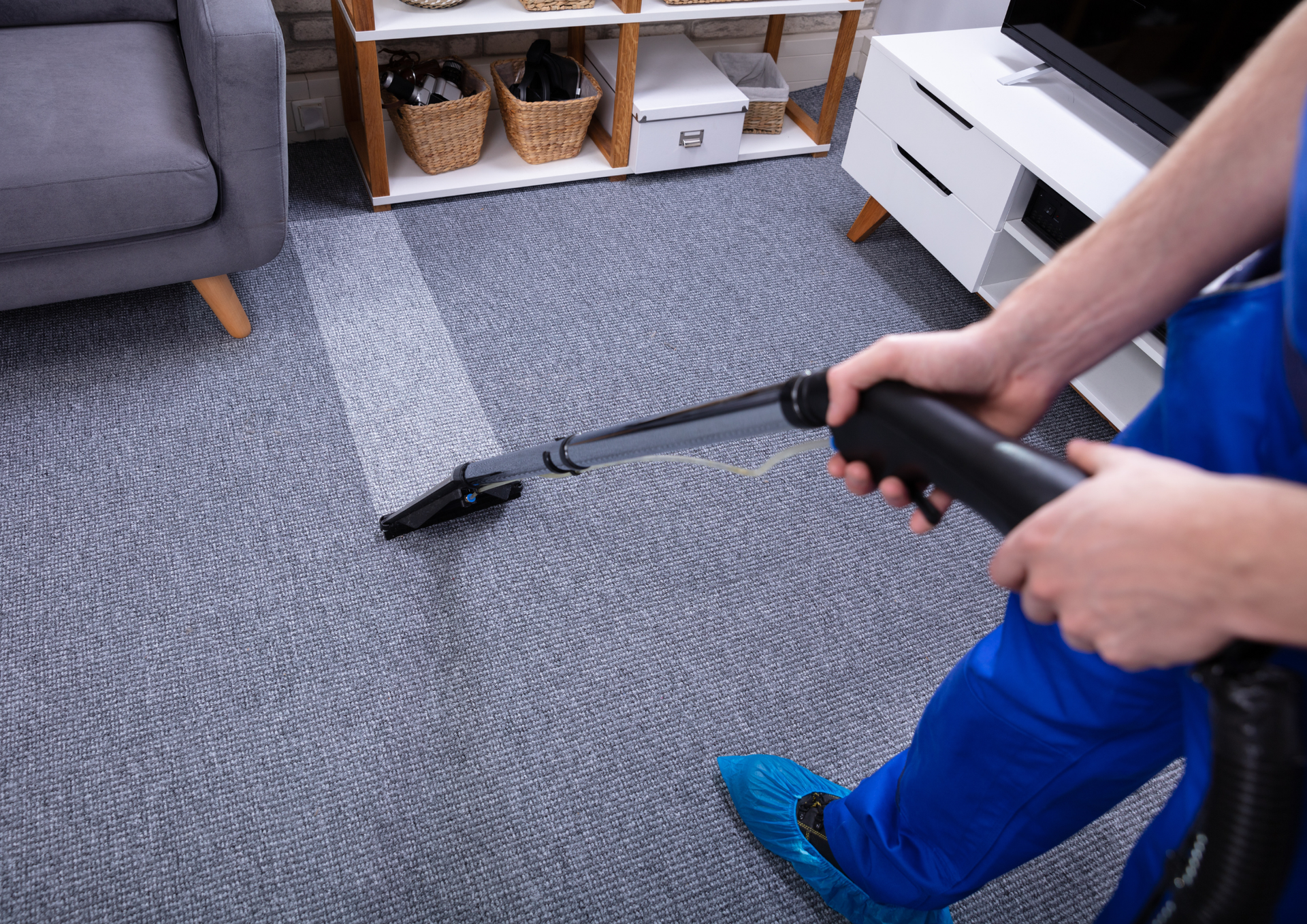 Person in blue cleaning a gray carpet with a vacuum, leaving a lighter cleaned path.
