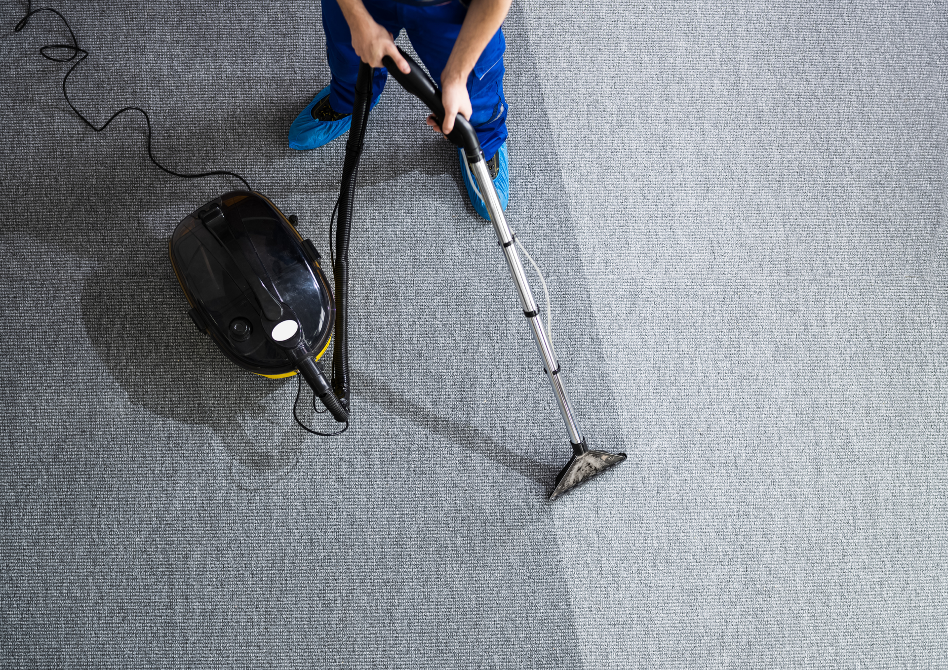 Person vacuuming gray carpet with a black machine and wand; cleaning in progress.