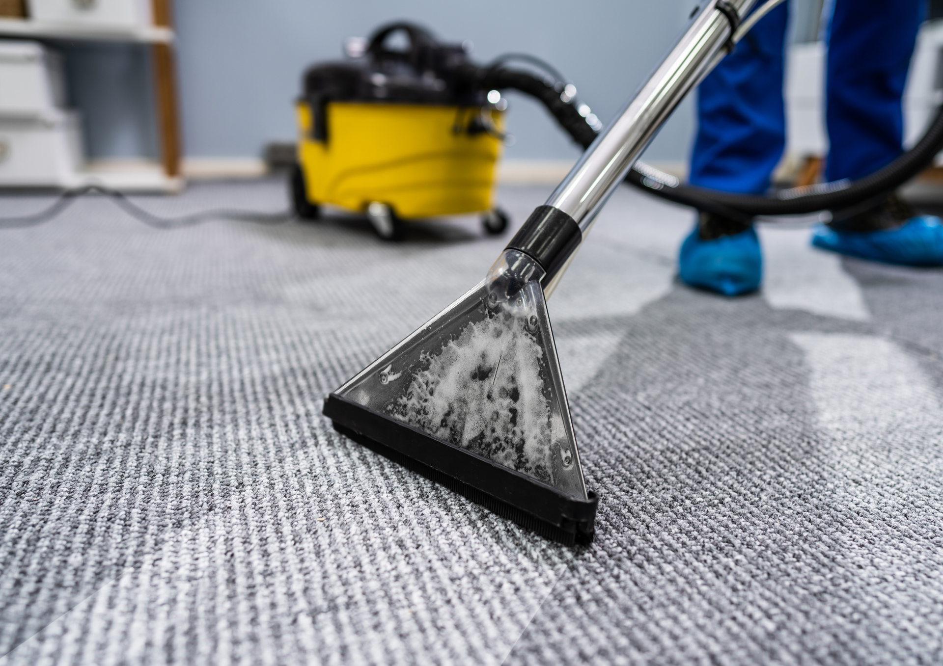Carpet being cleaned with a yellow vacuum cleaner. Person in blue shoe covers.