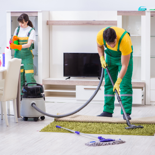 Two people cleaning a living room: one vacuums rug, the other wipes a cabinet. Both wear green overalls and gloves.