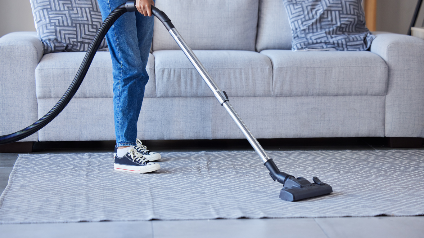 Person vacuuming a patterned rug in a living room, with a gray couch and pillows in the background.