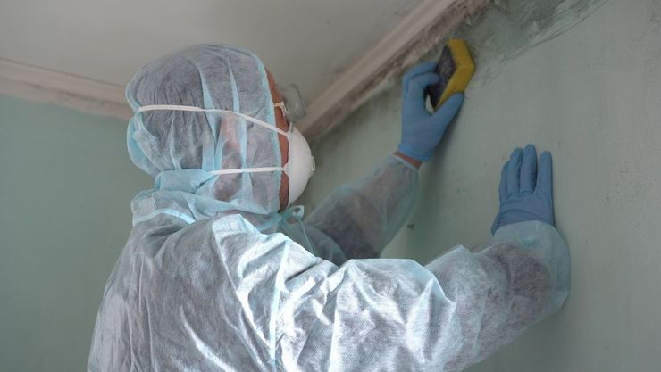 Worker in protective suit scrapes mold or damage from a ceiling corner with a tool.