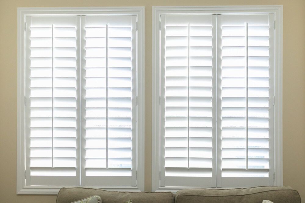 A Living Room With Two Windows With White Shutters And A Couch — Mid Coast Flooring & Blinds In Wingham, NSW