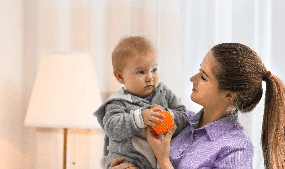 Woman holding a baby in a bright room, both looking at an orange toy.