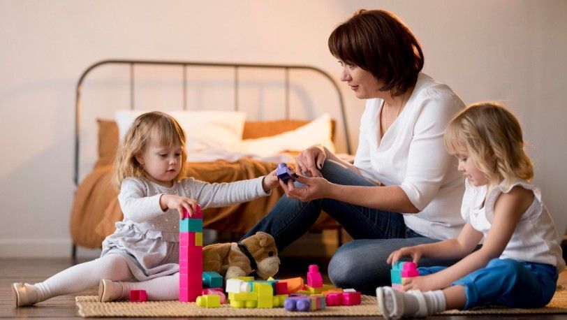 Woman playing with two children on a bed, surrounded by colorful toys in a cozy bedroom.
