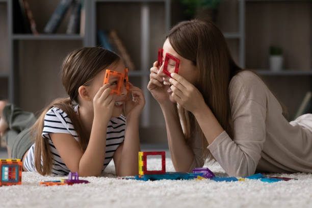 Two children lie on a rug, smiling and holding colorful toy glasses in a playroom.