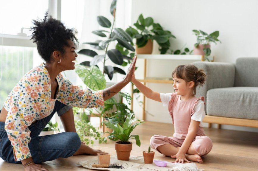 Adult and child high-five on a living room floor, with plants, books, and a gray sofa in the background