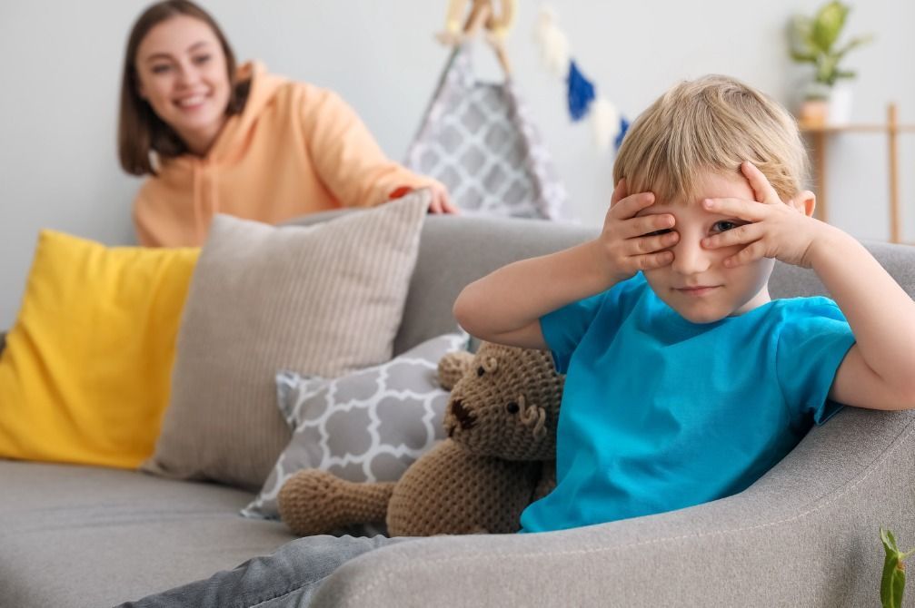 Child covering eyes on couch while smiling adult sits behind him in a bright living room