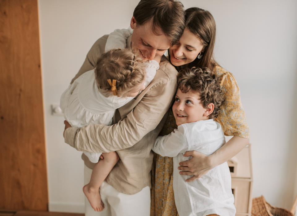 Family hugging together indoors, smiling in a warm embrace near a window.