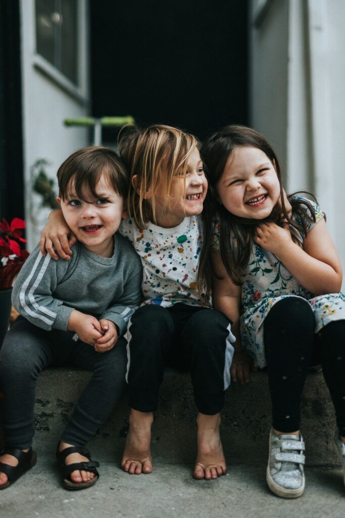 Three smiling children sitting on a doorstep, one leaning in for a hug