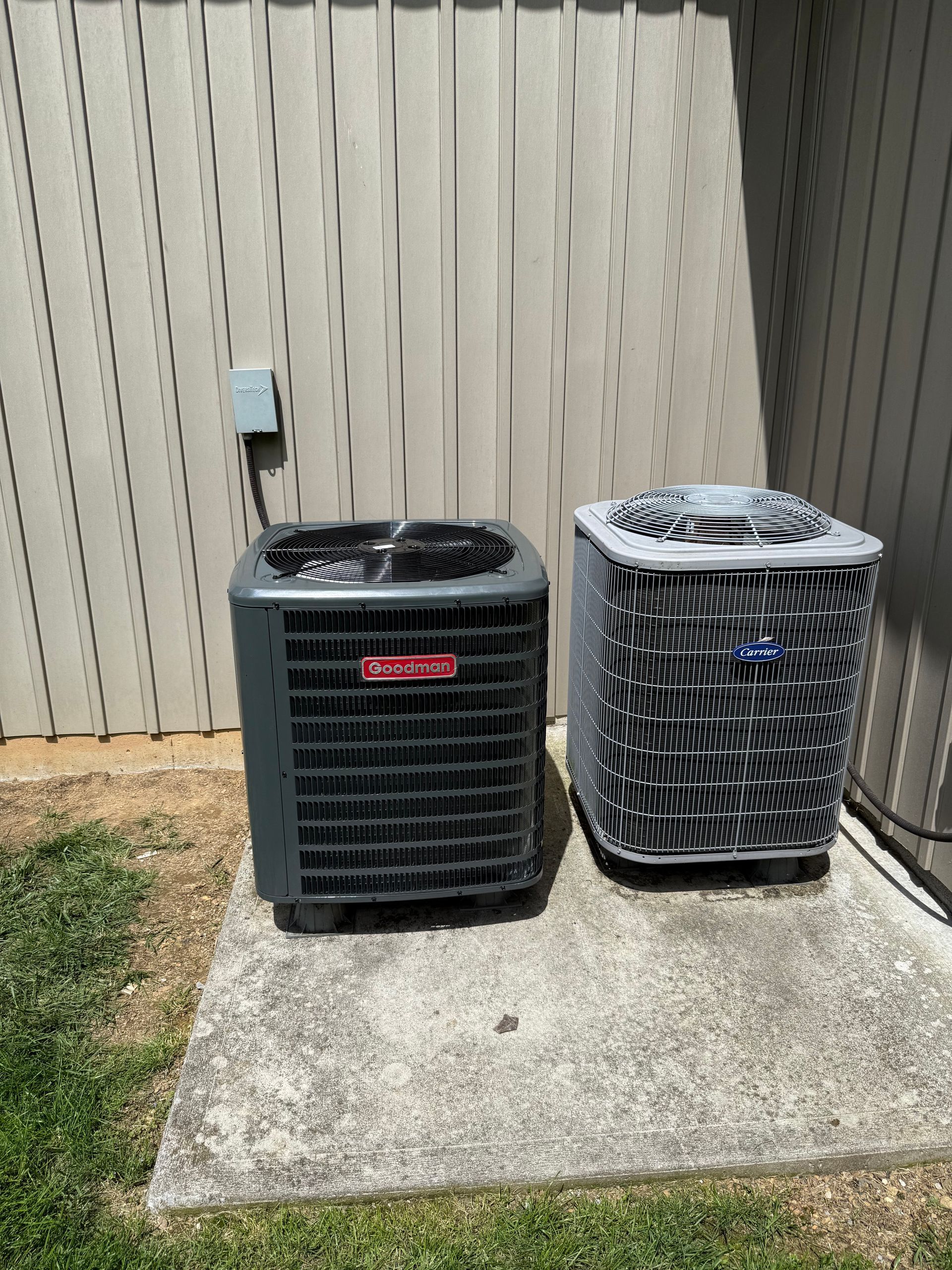 Two air conditioners are sitting on a sidewalk in front of a fence.