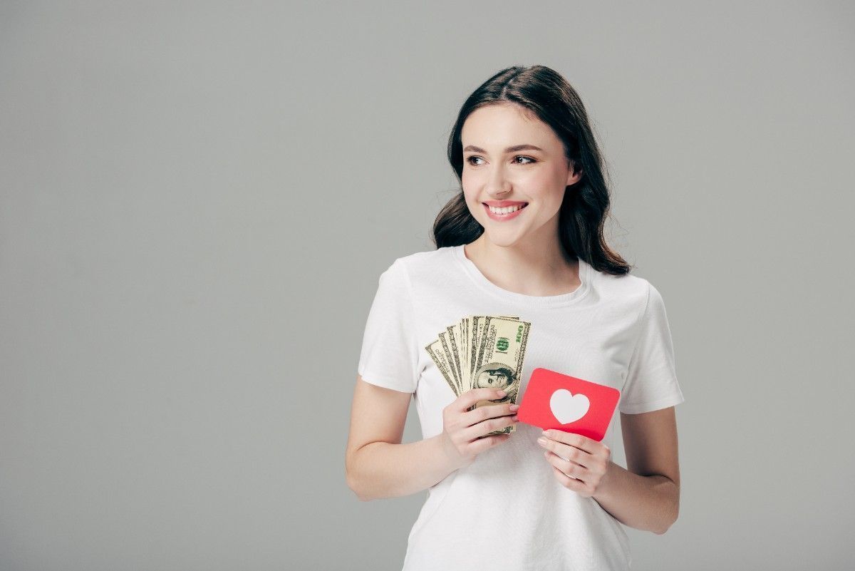 Woman smiling holding money and red card with heart symbol representing dating coaching services and relationship investment