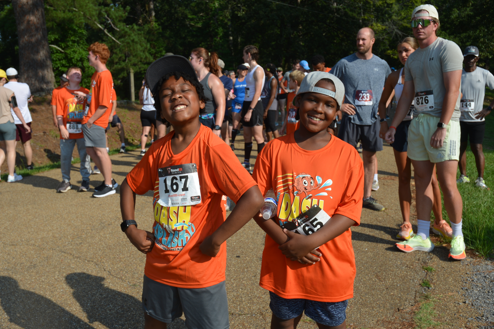 Two young boys wearing orange shirts are standing next to each other on a dirt road.