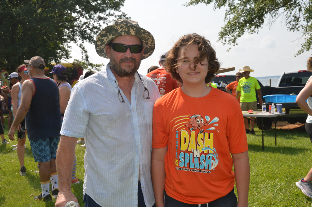 A man and a boy are posing for a picture in a park . the boy is wearing an orange dash splash shirt.