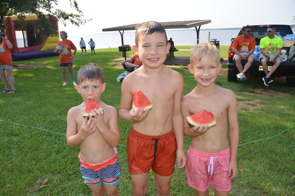 Three young boys are standing in the grass eating watermelon.
