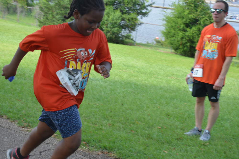 A boy in an orange shirt is running in a park while a man watches.