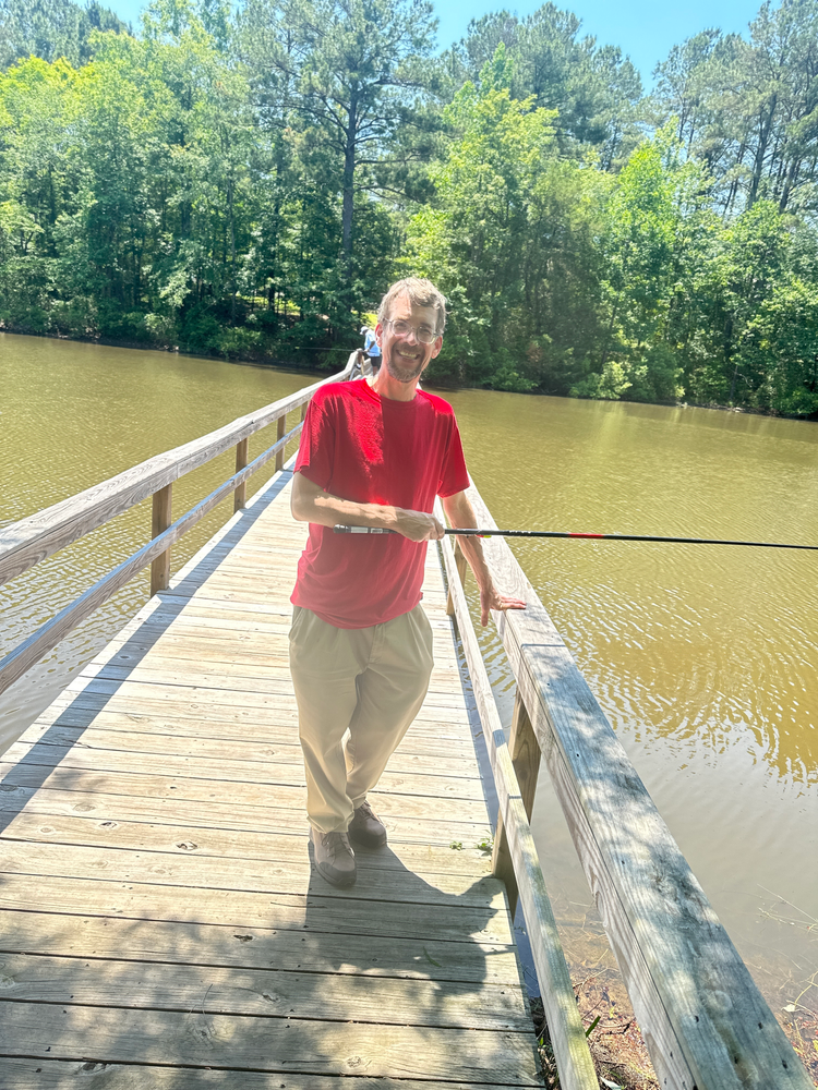 A man is standing on a wooden bridge over a lake holding a fishing rod.