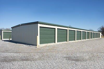 Row of green storage unit garages beside a metal building on a gravel lot under a clear blue sky