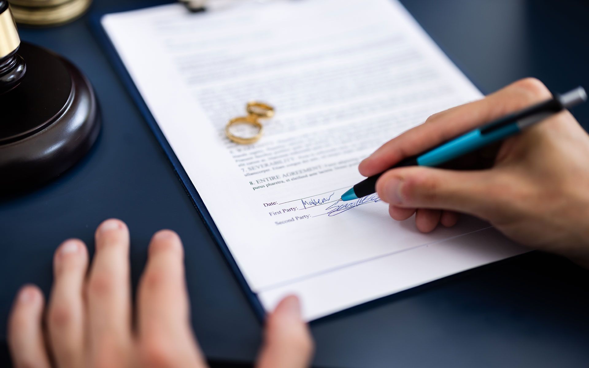 Person signing divorce papers, with wedding rings and gavel on a desk.