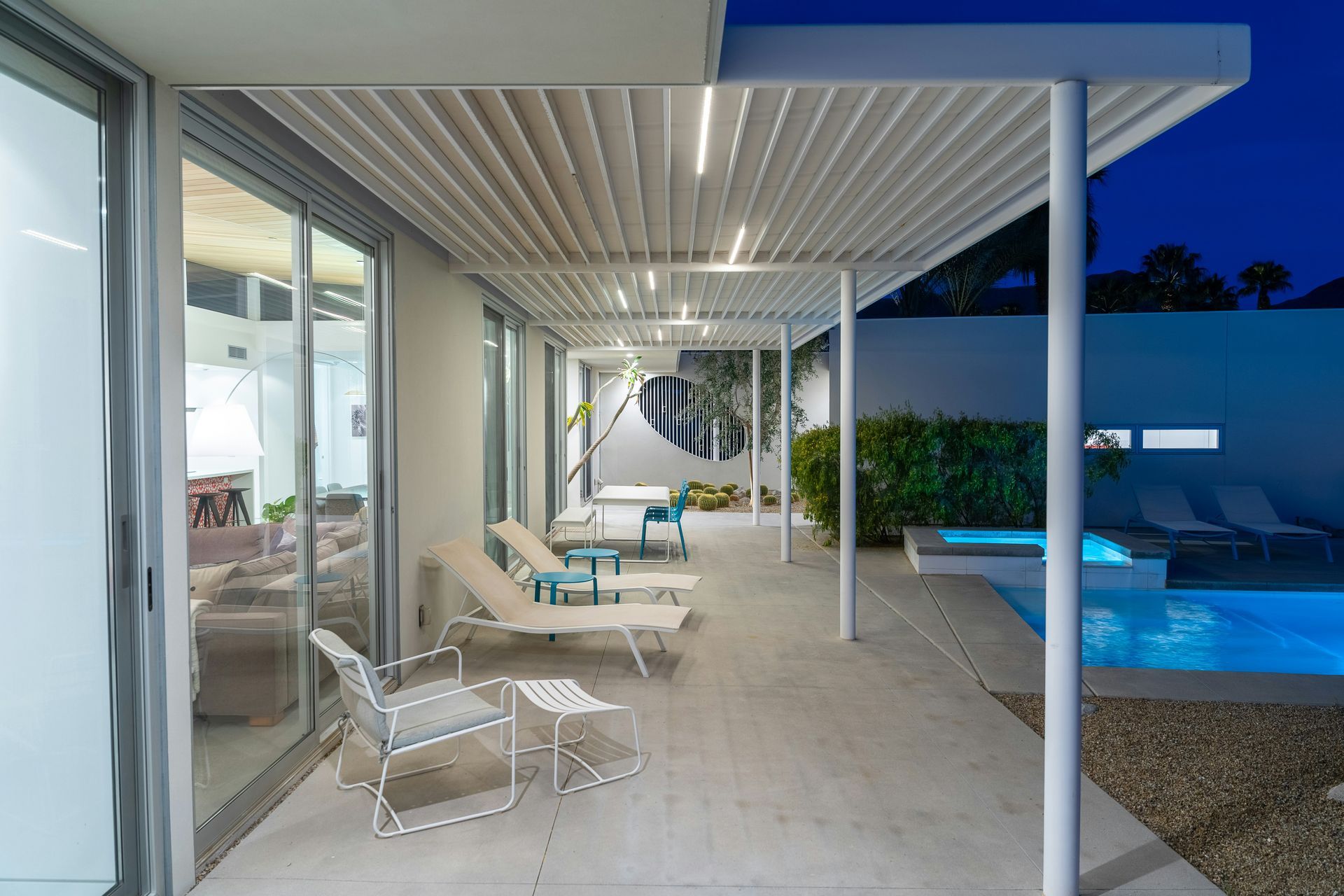 Covered patio with white chairs beside a lit swimming pool at dusk