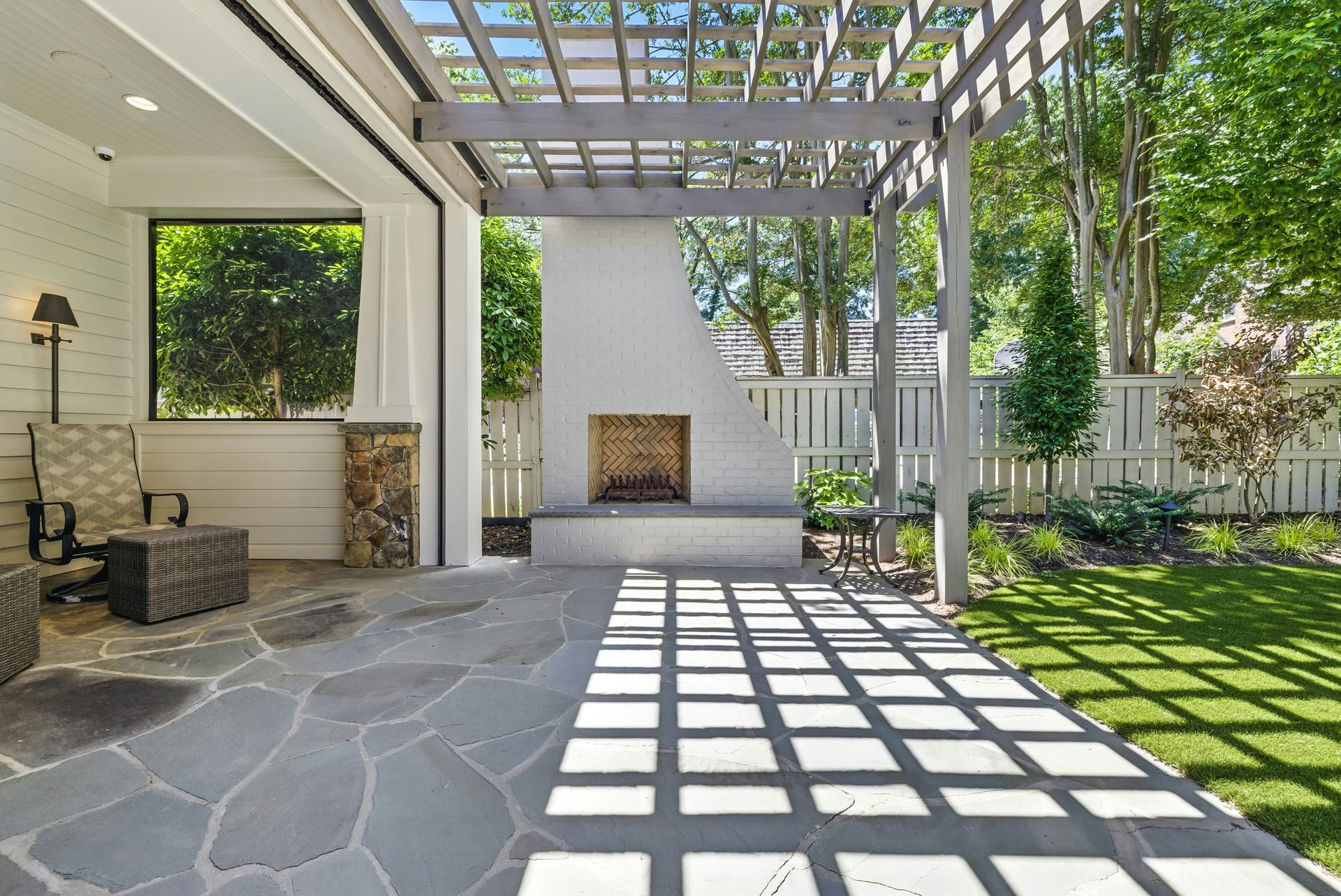 Covered stone patio with pergola, fireplace, and sunlit lattice shadows in a landscaped backyard