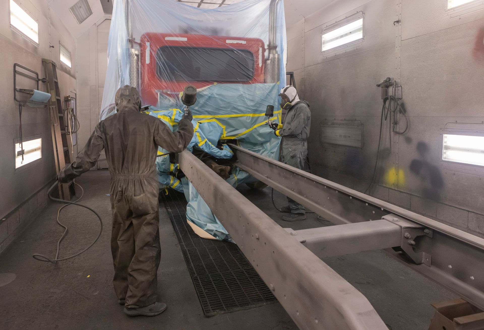 Two workers spray painting a truck frame in a paint booth.