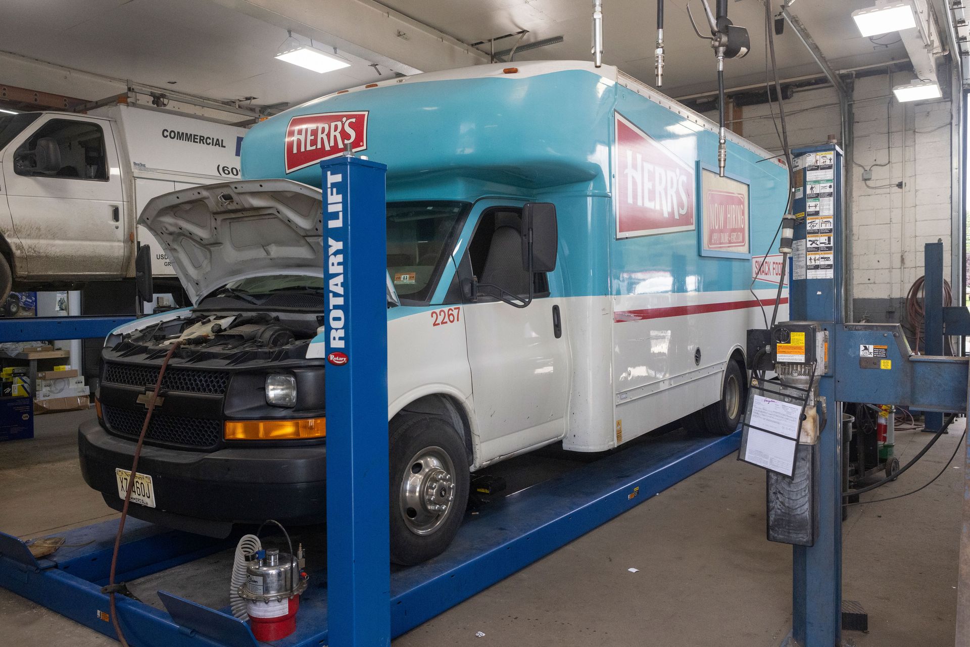An ice cream truck on a car lift in a repair shop with its hood open, light blue and white color scheme.