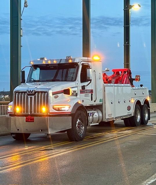 White tow truck with flashing lights on a bridge at dusk.