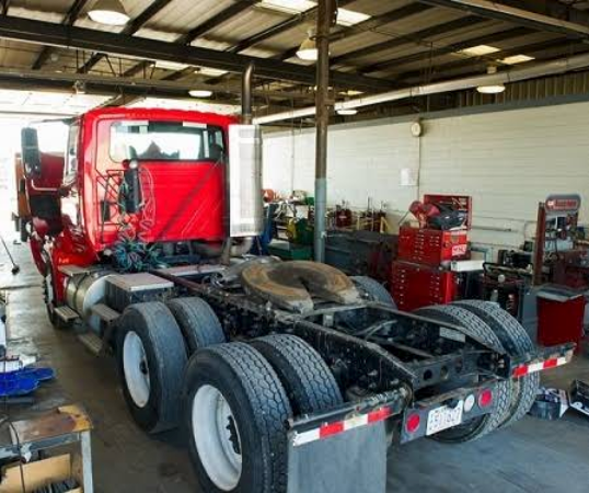 Red semi-truck in a repair shop, rear view. White tires, metal frame, various tools in background.