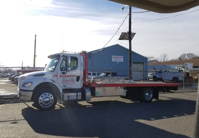 White tow truck parked in front of a blue building with a flatbed. Sunny day.