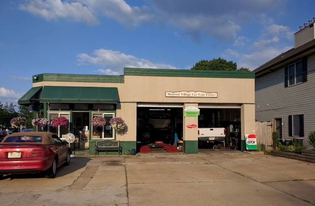 Small auto repair shop with open garage doors. Red car parked outside. Beige building with green accents.