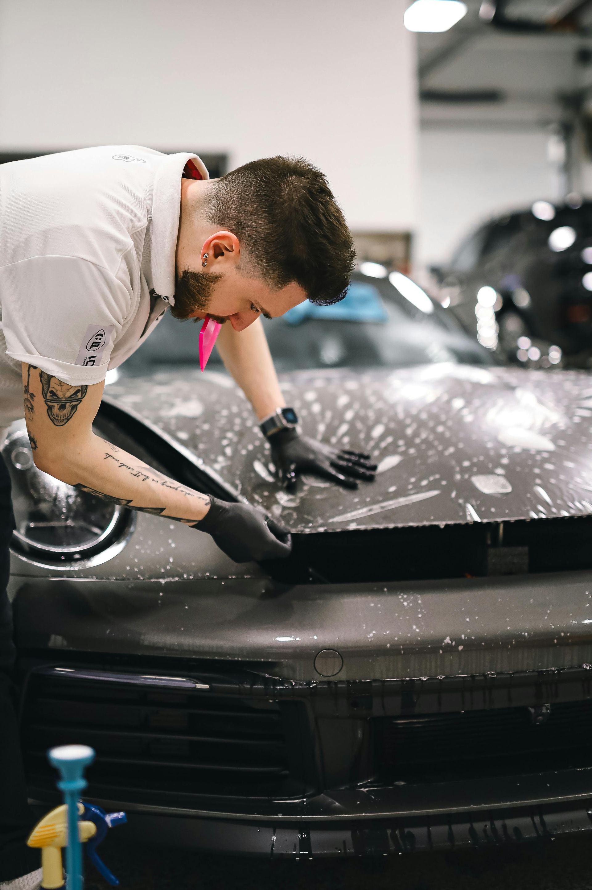Man applying protective film to a car's hood, inside a garage. He wears gloves.