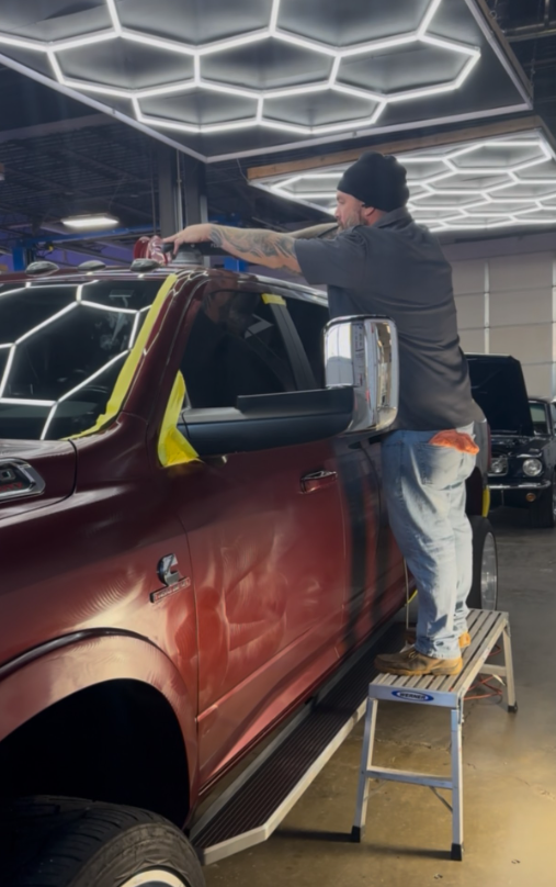 A person polishes a red pickup truck, standing on a step stool in a brightly lit shop.