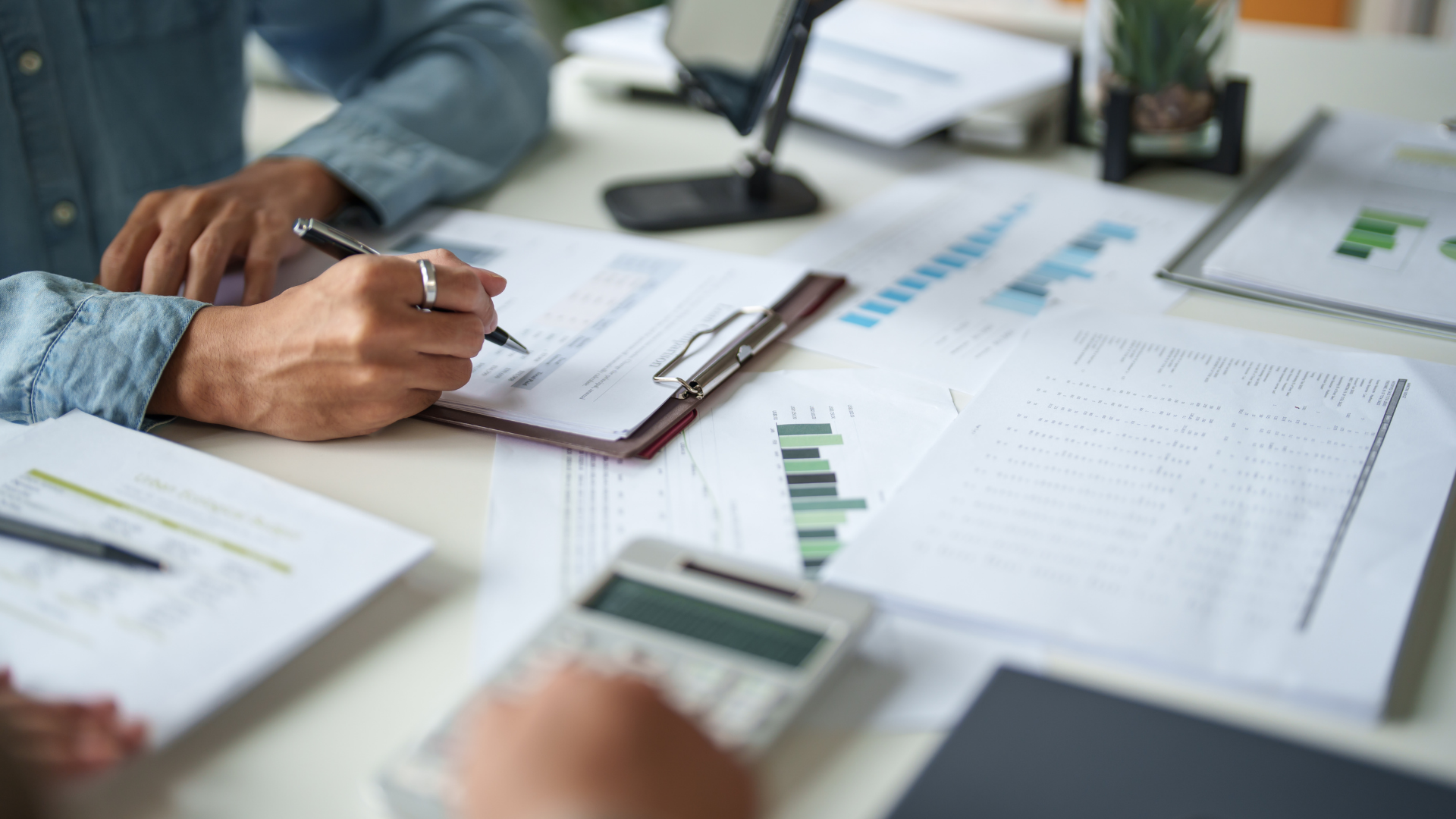 Hands taking notes on a clipboard beside printed charts and graphs in an office meeting.