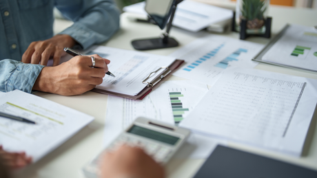 Hands reviewing financial charts and a clipboard at a desk with papers, graphs, and a laptop screen