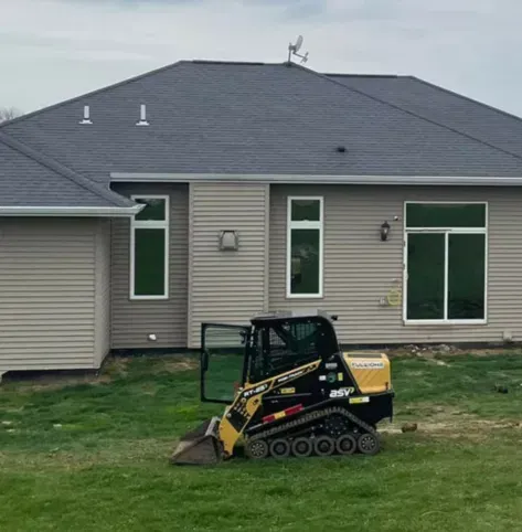Small yellow-and-black skid steer on grass in front of a gray house under cloudy skies