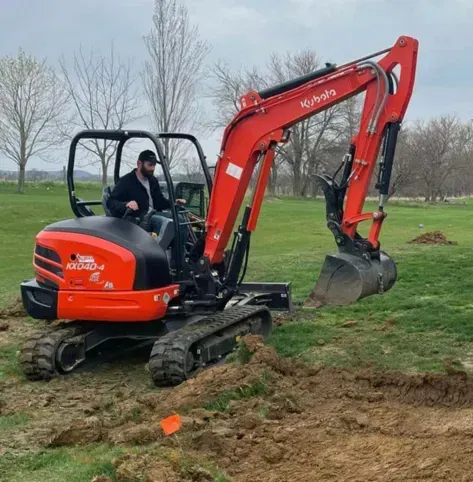 Orange excavator with operator digging soil in a grassy field with bare trees