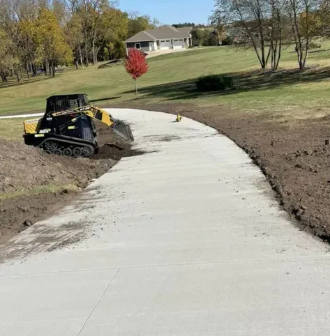 Concrete driveway under construction with a skid steer grading dirt beside a grassy hill.