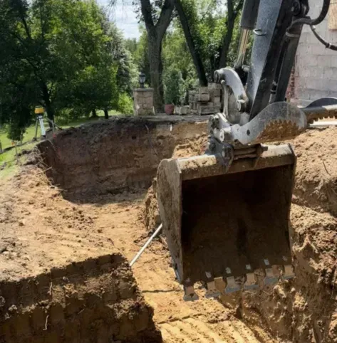 Excavator digging a large round pit in a wooded yard beside a building