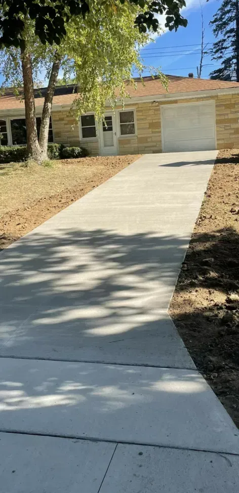 Concrete driveway leading to a single-story house with a garage and trees casting shade
