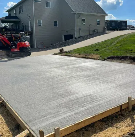 Fresh concrete slab in a residential driveway beside a house, with a red skid steer nearby.