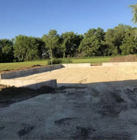 Empty concrete pool beside a grassy field and trees under a clear blue sky