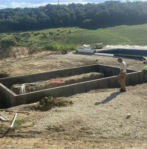 Construction site with a man standing by a large concrete foundation in a grassy field.