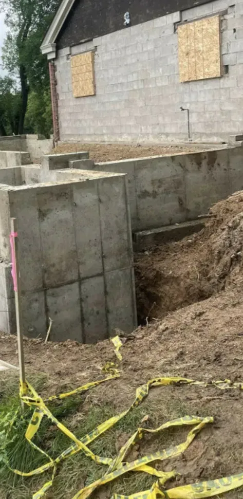 Foundation excavation beside a house with concrete walls, dirt trench, and yellow caution tape