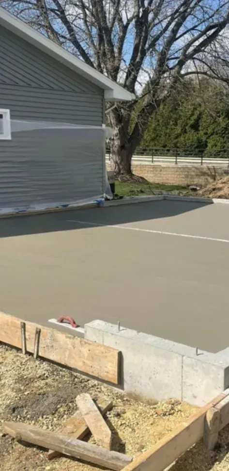 Concrete foundation slab beside a gray house, with wooden forms and a tree in the yard