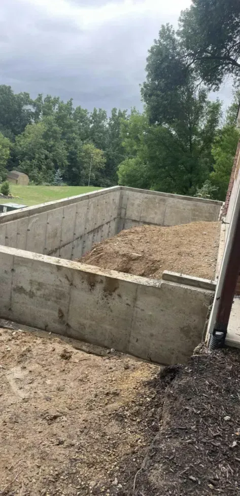 Concrete foundation walls for a small structure on a dirt lot, with trees and cloudy sky in the background