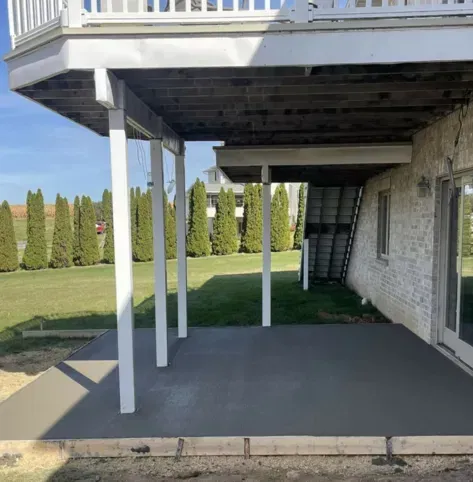 Under-deck patio with white support posts, black concrete floor, and brick house wall beside a lawn.